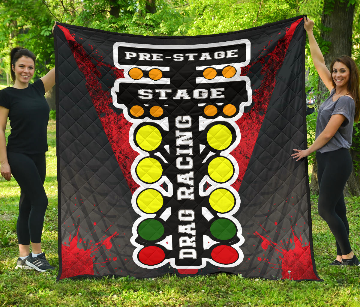 Two women holding a red-and-black drag racing quilt outdoors, showing starting-tree lights, tire-track artwork, and diamond stitching.