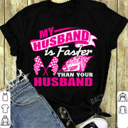 Woman sitting in the grandstands at a race track wearing a dark gray v-neck t-shirt with a pink and white graphic that reads "My Husband Is Faster Than Your Husband" alongside a race car and checkered flags.
