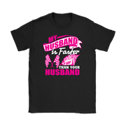 Woman sitting in the grandstands at a race track wearing a dark gray v-neck t-shirt with a pink and white graphic that reads "My Husband Is Faster Than Your Husband" alongside a race car and checkered flags.