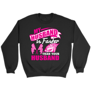 Woman sitting in the grandstands at a race track wearing a dark gray v-neck t-shirt with a pink and white graphic that reads "My Husband Is Faster Than Your Husband" alongside a race car and checkered flags.