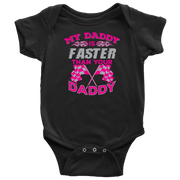 A proud, smiling racing dad in a fire suit holding his baby, who is wearing a cute "My Daddy Is Faster Than Your Daddy" bodysuit, standing confidently next to a colorful race car in the pit area.