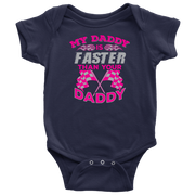 A proud, smiling racing dad in a fire suit holding his baby, who is wearing a cute "My Daddy Is Faster Than Your Daddy" bodysuit, standing confidently next to a colorful race car in the pit area.
