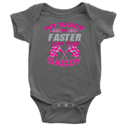 A proud, smiling racing dad in a fire suit holding his baby, who is wearing a cute "My Daddy Is Faster Than Your Daddy" bodysuit, standing confidently next to a colorful race car in the pit area.